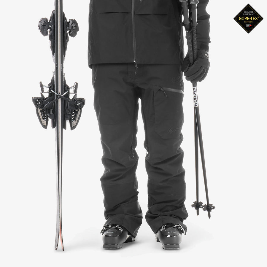 A person wearing black ski pants stands against a light gray studio background, holding ski poles and next to a pair of skis. A black Gore-Tex logo patch is visible in the top right corner.