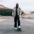 A man stands on a skateboard on an outdoor paved road, wearing a beige and black patterned jacket, black pants, and white sneakers, with a white cap. The background shows a cloudy sky, dunes, and low buildings.