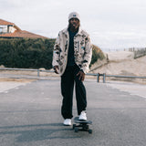 A man stands on a skateboard on an outdoor paved road, wearing a beige and black patterned jacket, black pants, and white sneakers, with a white cap. The background shows a cloudy sky, dunes, and low buildings.