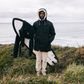 A man wearing a black hooded puffer jacket and beige pants stands on a grassy cliffside overlooking the ocean, holding a black and white surfboard under his arm.