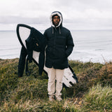 A man wearing a black hooded puffer jacket and beige pants stands on a grassy cliffside overlooking the ocean, holding a black and white surfboard under his arm.