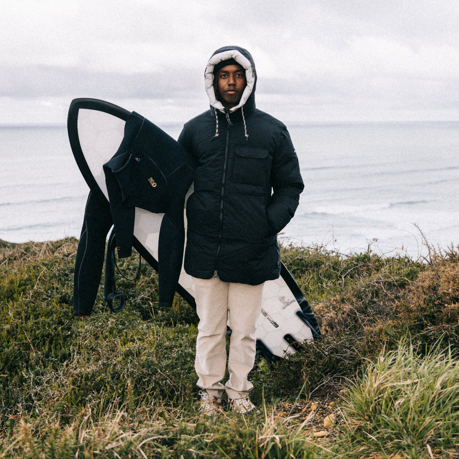 A man wearing a black hooded puffer jacket and beige pants stands on a grassy cliffside overlooking the ocean, holding a black and white surfboard under his arm.