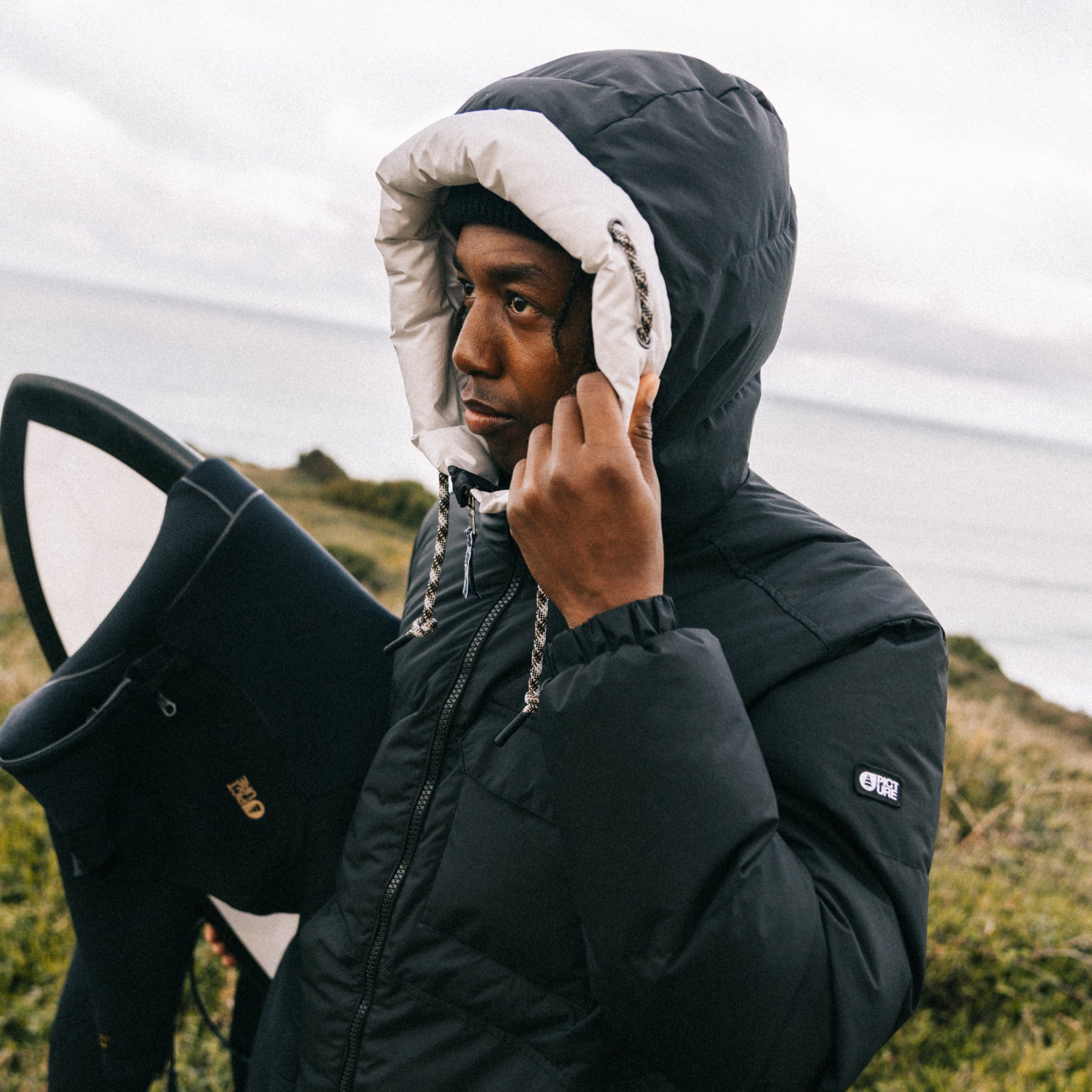 A man wearing a black hooded puffer jacket with a white fleece-lined hood stands outdoors near the ocean, holding a surfboard.