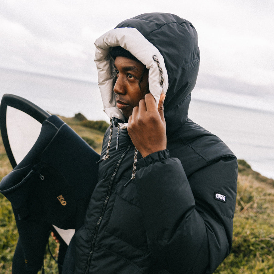 A man wearing a black hooded puffer jacket with a white fleece-lined hood stands outdoors near the ocean, holding a surfboard.