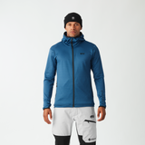 A man modeling a blue technical zip-up jacket and white, black, and gray ski pants against a plain light gray studio background.