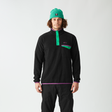A man stands against a light gray studio background wearing a black quarter-zip fleece with a bright green zipper and chest logo, black pants, and a green beanie.
