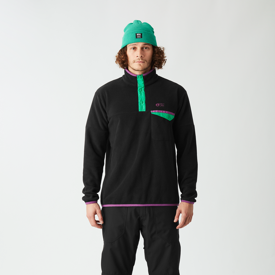 A man stands against a light gray studio background wearing a black quarter-zip fleece with a bright green zipper and chest logo, black pants, and a green beanie.