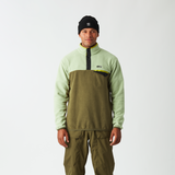 A man stands against a light gray studio background wearing a light sage green and olive green quarter-zip pullover with a black zipper and chest logo, paired with olive green snow pants and a black beanie.