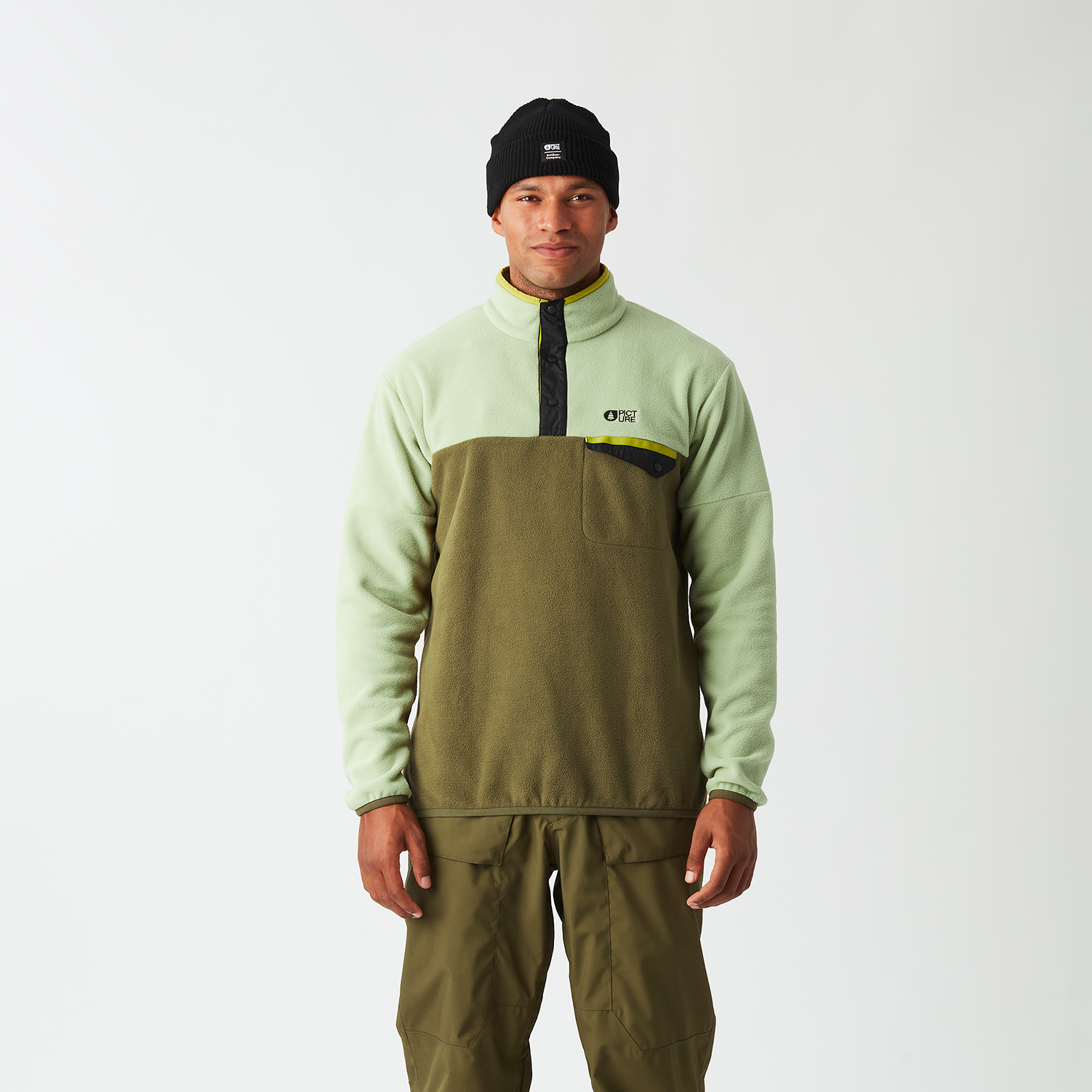 A man stands against a light gray studio background wearing a light sage green and olive green quarter-zip pullover with a black zipper and chest logo, paired with olive green snow pants and a black beanie.