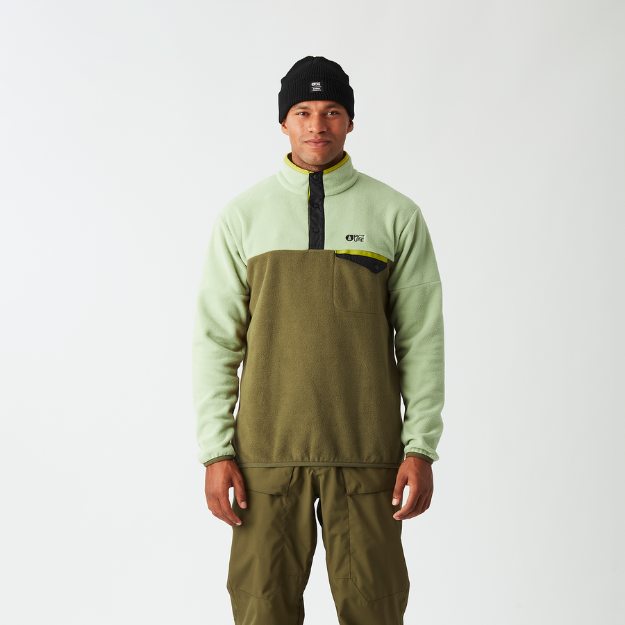 A man stands against a light gray studio background wearing a light sage green and olive green quarter-zip pullover with a black zipper and chest logo, paired with olive green snow pants and a black beanie.