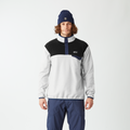 A man wearing a color-blocked quarter-zip pullover jacket in white, black, and navy, paired with navy pants, a navy beanie, and gray sneakers, standing against a plain white studio background.