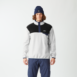 A man wearing a color-blocked quarter-zip pullover jacket in white, black, and navy, paired with navy pants, a navy beanie, and gray sneakers, standing against a plain white studio background.