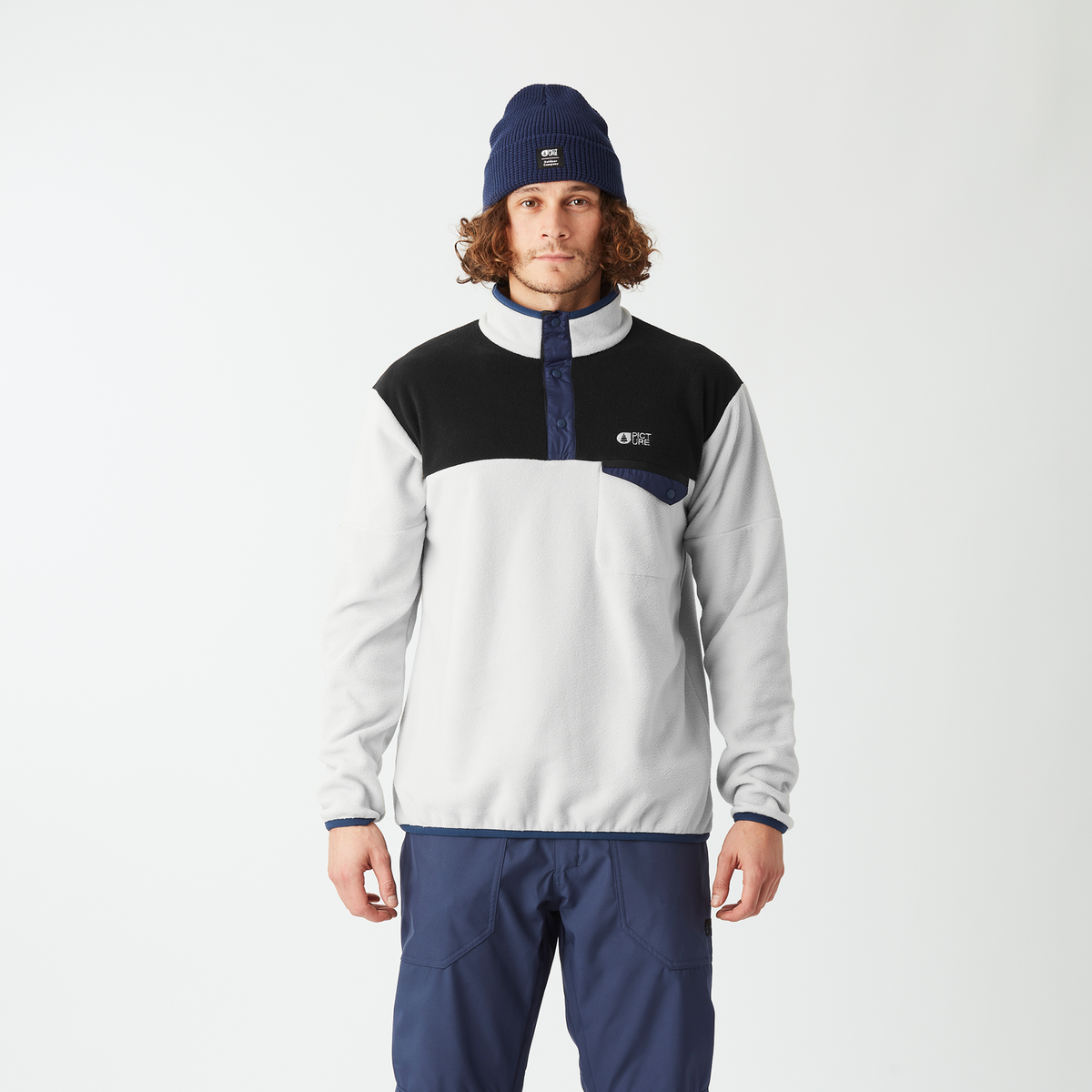 A man wearing a color-blocked quarter-zip pullover jacket in white, black, and navy, paired with navy pants, a navy beanie, and gray sneakers, standing against a plain white studio background.