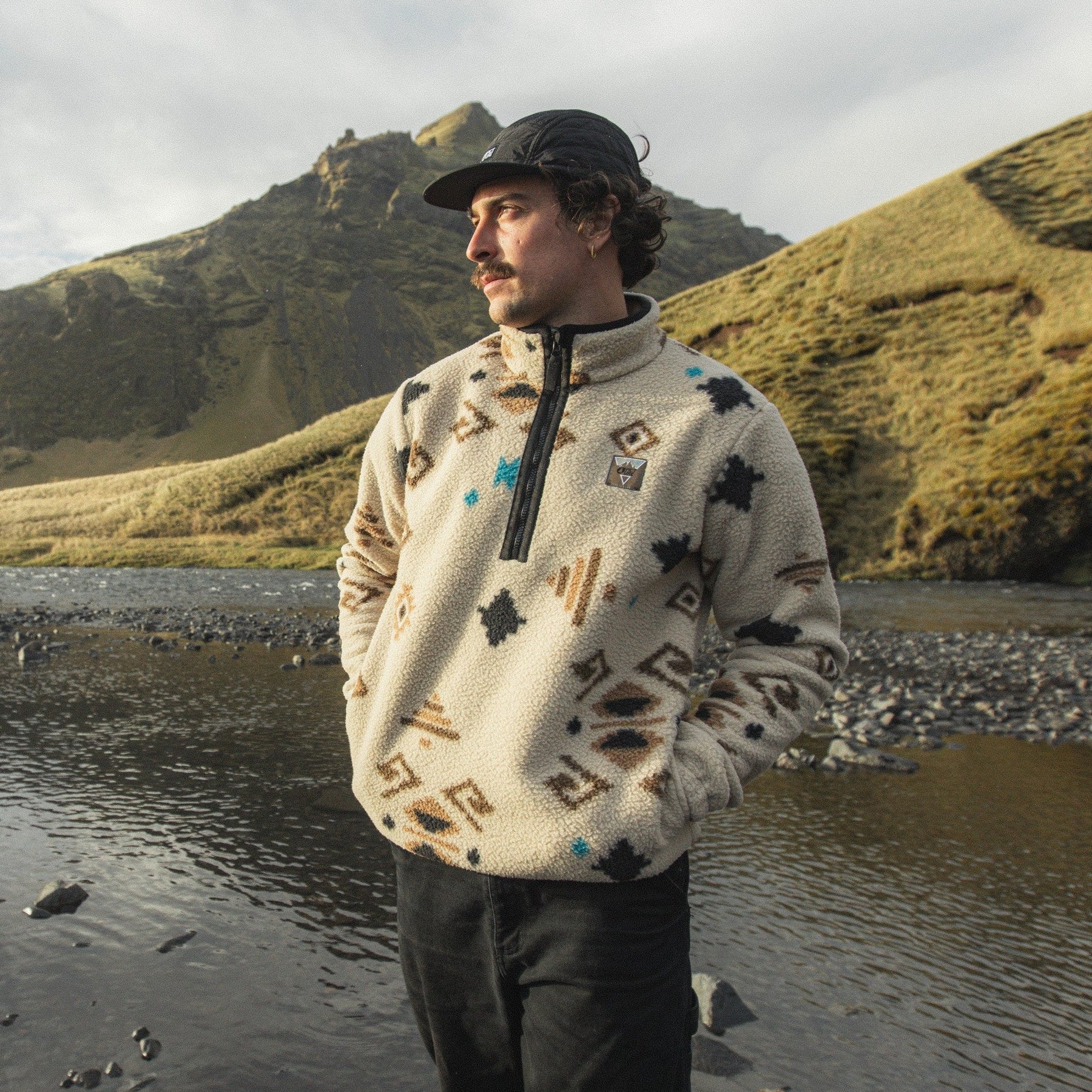 A man wearing a cream fleece pullover with a geometric pattern, black pants, and a black cap stands on a rocky riverbank with grassy mountains in the background.