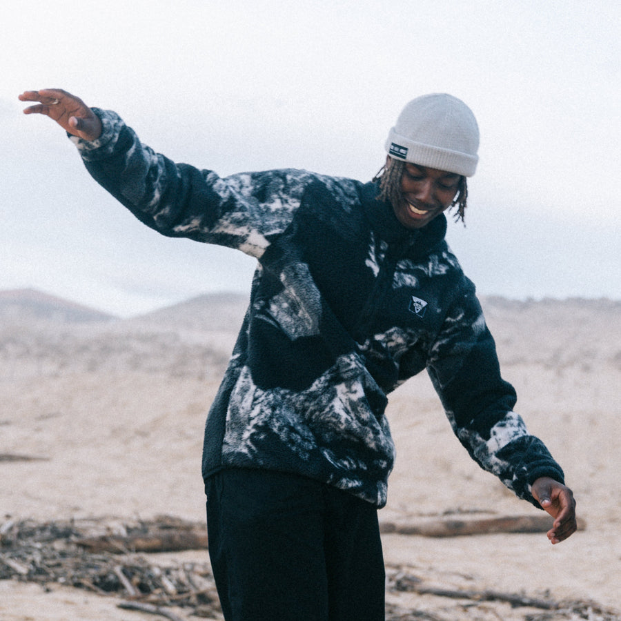 A person walking outdoors on a sandy beach dune wearing a black and gray patterned jacket, black pants, and a white beanie.