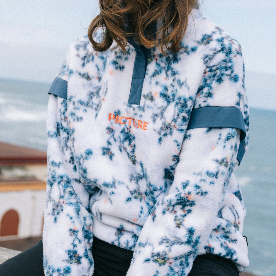 A woman wearing a white fleece pullover with a blue floral pattern, dark blue trim, and an orange 'LIFECURE' logo, sitting outdoors with the ocean and cloudy sky in the background.