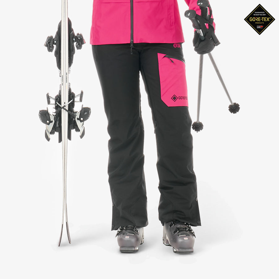 A person wearing black ski pants with bright pink accents and a matching pink jacket, standing on a white studio background with skis and ski poles. A black and gold Gore-Tex logo badge appears in the top right corner.