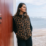 A woman wearing a brown and black leopard print zip-up vest over a black long-sleeve top, leaning against a rustic red wooden door with a stone path and the ocean in the background.