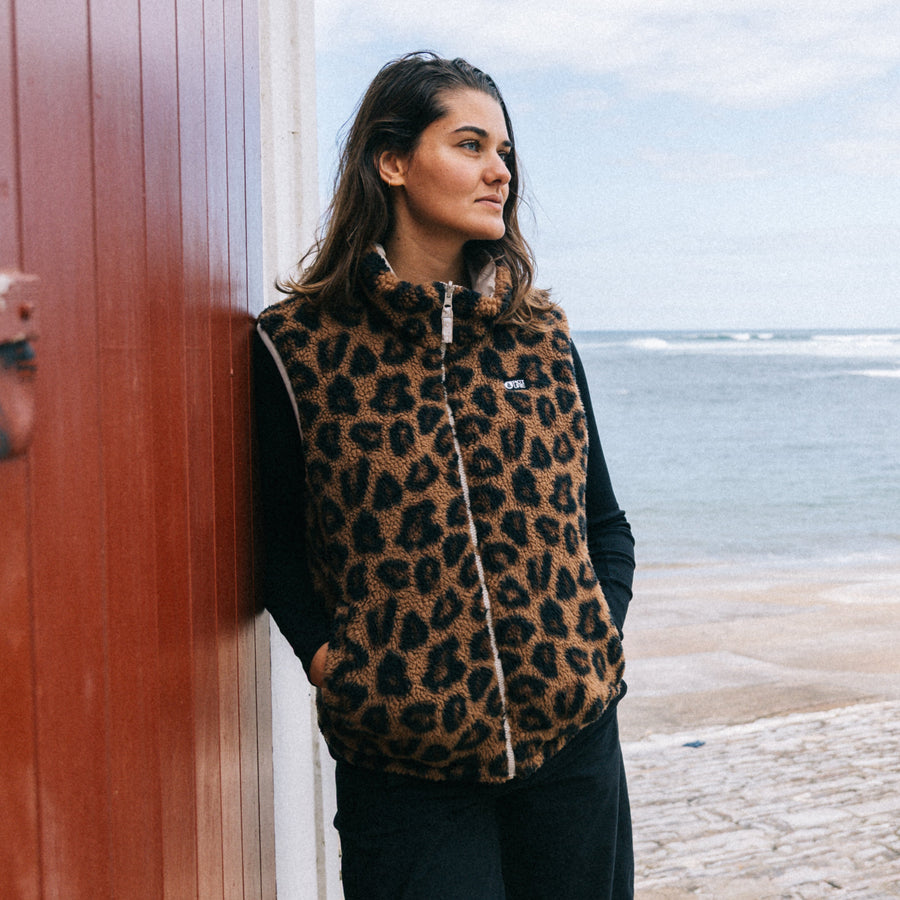 A woman wearing a brown and black leopard print zip-up vest over a black long-sleeve top, leaning against a rustic red wooden door with a stone path and the ocean in the background.